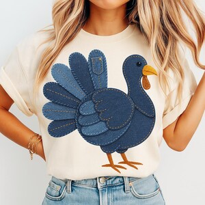 Könnte beinhalten: Cremefarbenes T-Shirt mit einem Denim-Truthahn-Design. Der Truthahn hat einen blauen Körper, einen gelben Schnabel und eine braune Wamme. Die Schwanzfedern des Truthahns bestehen aus Jeans-Patches mit sichtbaren Nähten. Das Shirt ist ein lässiger Stil mit kurzen Ärmeln.