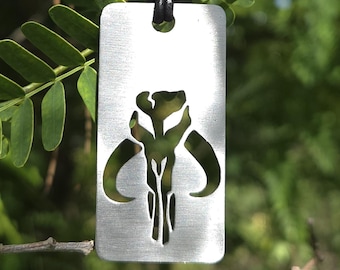 May include: A silver-colored rectangular pendant necklace with a black cord. The pendant features a cut-out design of a stylized skull with horns. The background is a blurred view of green leaves and branches.