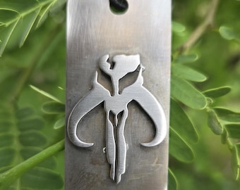 May include: A rectangular silver pendant featuring a Mandalorian skull symbol. The pendant hangs from a black leather cord. The background is a blurred view of green leaves.
