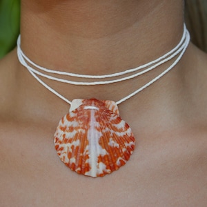 May include: A layered white cord necklace featuring a large, orange and white seashell pendant. The shell has a detailed pattern and hangs from the necklace, creating a beachy, natural aesthetic. The necklace is worn around the neck.