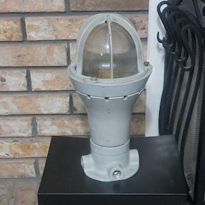 May include: A vintage, industrial-style light fixture with a gray, cylindrical base and a clear, domed glass cover. The fixture has a cage-like structure around the glass and is mounted on a black surface. The background features a brick wall.