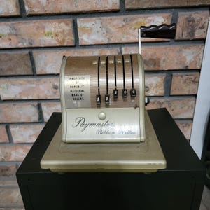 May include: An antique Paymaster Ribbon Writer, a vintage machine with a beige and silver finish. The machine has a black handle and a series of dollar and cent indicators. The text "PROPERTY OF REPUBLIC NATIONAL BANK OF DALLAS" is visible.