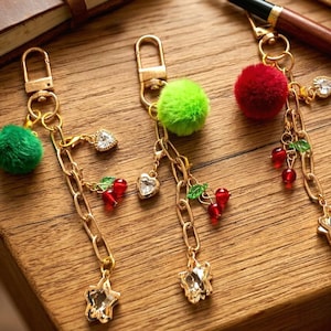 May include: Three gold-toned keychains with decorative charms. Each keychain features a fluffy pom-pom in green or red, a heart-shaped charm, and a star-shaped charm. Additional charms include red cherry clusters with green leaves.