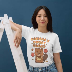 May include: White t-shirt with the text "CANADA'S SWEET TREAT" above a cartoon beaver holding a pastry. The beaver is brown with a large tail and is surrounded by maple leaves. The shirt is worn by a person.