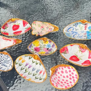 May include: A collection of hand-painted seashells with various designs. Some shells feature strawberries, floral patterns, and lobster illustrations. Others have a blue and white floral design with the text "Cape Cod".