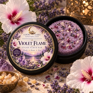 Könnte beinhalten: Eine runde schwarze Dose mit einem violetten Zuckerpeeling-Stück, beschriftet als Violet Flame Sugar Scrub Bar von Loon Lake Apothecary. Der Deckel zeigt einen Halbmond, Amethystkristalle und Hibiskusblüten. Das Stück wiegt 57 Gramm.
