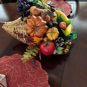 May include: A woven cornucopia centerpiece filled with artificial autumn harvest items including a large orange pumpkin, a smaller yellow pumpkin, a red apple, a yellow banana, purple grapes, a pinecone, and various green and brown leaves.