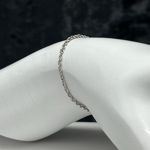 May include: A silver-colored rope chain bracelet. The bracelet is draped over a white surface, with a dark background. The chain has a twisted rope design, and the bracelet appears to be made of metal.