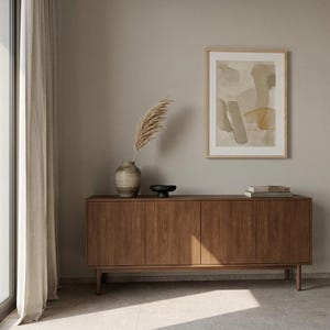 Peut inclure: Un buffet rectangulaire en bois à quatre portes repose sur quatre pieds. Sur le dessus se trouvent un vase en céramique avec un plumeau d'herbe de la pampa séchée et un petit bol noir. Une impression abstraite encadrée est fixée au mur.