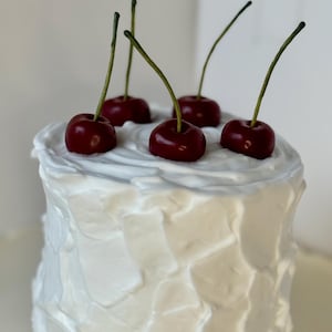 May include: A white frosted cake topped with five dark red cherries with green stems. The cake has a textured, layered frosting design. The cherries are glossy and appear fresh.