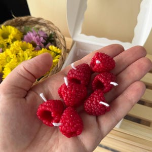 May include: A hand holds several bright red raspberry-shaped candles with white wicks. The candles have a textured surface, mimicking the appearance of real raspberries. A basket of yellow and purple flowers and a white box are in the background.