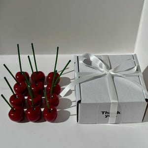 May include: A collection of glossy red cherry ornaments with green stems are arranged near a white gift box tied with a white ribbon. The box has the words "Thank you" printed on it.