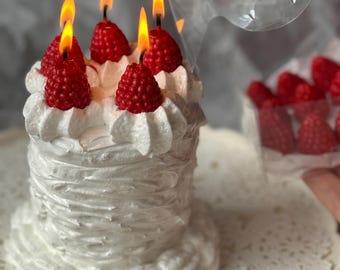 May include: A white, frosted candle shaped like a cake, adorned with red raspberry-shaped candles with lit flames. The candle sits on a white, lace-patterned doily. A clear plastic container holds additional raspberry-shaped candles.