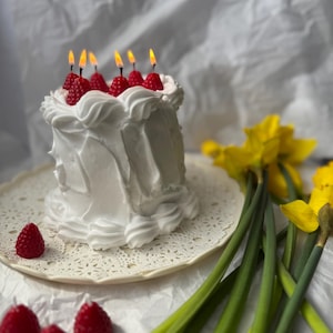 May include: A white cake-shaped candle with red raspberry-shaped candles on top, each with a lit wick. The candle sits on a white lace doily, with additional raspberry candles nearby. Yellow daffodils with green stems are in the background.