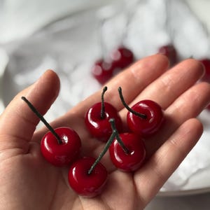 May include: Six glossy, bright red cherries with dark green stems are held in a hand. The cherries have a realistic appearance, with a shiny, reflective surface. The background is blurred, suggesting a shallow depth of field.