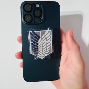 May include: A dark blue smartphone with a clear emblem featuring a stylized wing design in white, blue, and brown. The emblem is affixed to the phone's back. The phone has a multi-lens camera system.