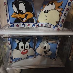 May include: Four square throw pillows featuring cartoon characters. The pillows have a blue background with Daffy Duck, Taz, Sylvester, and Bugs Bunny. Each pillow has a white border with colorful star and flower designs.