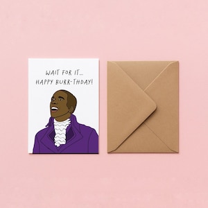 May include: A greeting card with a cartoon illustration of a man in a purple coat and white ruffled shirt. The card reads "WAIT FOR IT... HAPPY BURR-THDAY!" and is accompanied by a brown envelope, all set against a pink background.