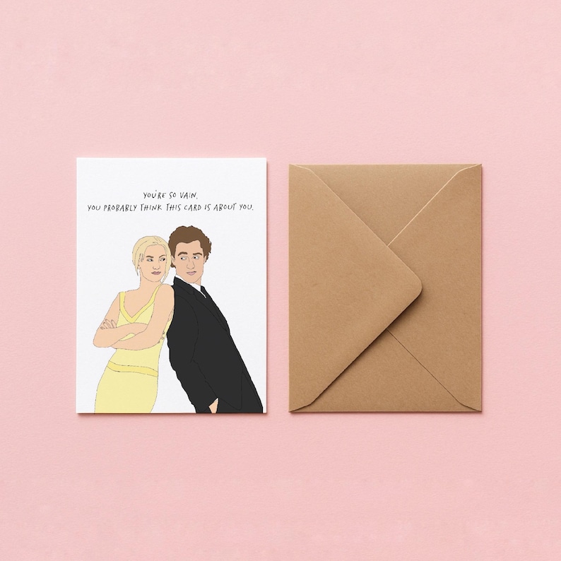 以下が含まれることがあります： 「You're so vain, you probably think this card is about you.」のテキストとカップルのイラストが描かれたユーモラスなグリーティングカード。茶色の封筒がピンク色の背景にカードの隣に置かれています。