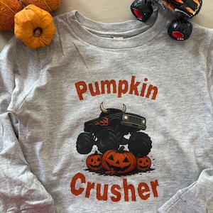 Puede incluir: Una camiseta gris de manga larga con las palabras "Pumpkin Crusher" en naranja. La camiseta presenta un gráfico de un camión monstruo negro con cuernos de toro y tres calabazas talladas. También hay calabazas decorativas naranjas y un camión monstruo de juguete.