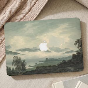 May include: Laptop case with a landscape painting depicting mountains, a lake, and trees in muted green and grey tones. The Apple logo is visible. The case is on a beige surface.