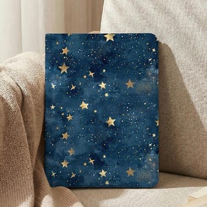 Puede incluir: Una funda rectangular azul oscuro con un diseño celestial. Presenta estrellas doradas y motas blancas sobre un fondo de estilo acuarela. La funda está sobre una superficie beige.