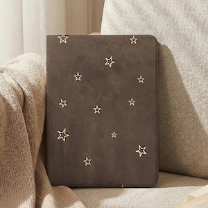 Op de afbeelding: Een rechthoekig donkerbruin notitieboek of tablethoes met een verspreid patroon van goudkleurige steromtrekken. De kaft heeft een gladde, matte textuur en afgeronde hoeken, wat zorgt voor een strak ontwerp zonder extra tekst of markeringen.