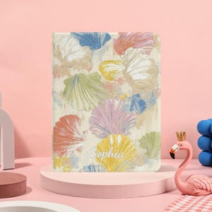 May include: A tablet case featuring a seashell design in pastel colors, including pink, yellow, blue, and green. The name "Sophia" is written in cursive on the front of the case. The case has a beige background.