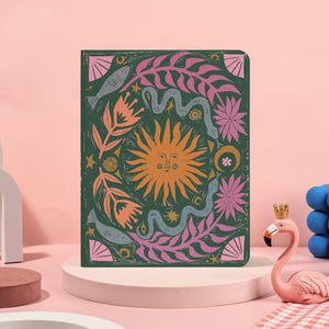 May include: A dark green journal with a colorful illustration featuring a smiling sun, snakes, flowers, and fish. The design is in shades of orange, pink, and teal. The journal is displayed on a pink pedestal with a flamingo statue in the foreground.