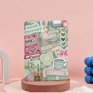 Könnte beinhalten: Eine dekorative Handyhülle mit Buchmotiv, die eine Collage aus Buchstapeln, einer Kaffeetasse, Herzen und Schleifen zeigt. Die Hülle trägt den Text "book therapy" und "BOOKS Because reality is meh". Der Hintergrund ist ein grün-weiß kariertes Muster.