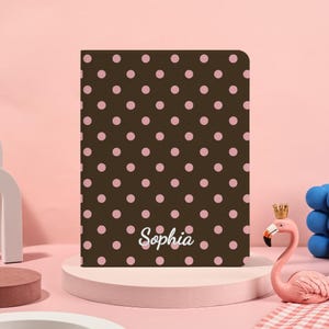 Op de afbeelding: Een bruin notitieboek met een roze stippenpatroon. Het woord "Sophia" staat in witte letters onderaan. Het notitieboek staat op een roze sokkel, met een roze flamingo en een blauwe bolsculptuur op de achtergrond.