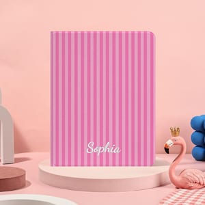 Peut inclure: Un carnet rose à rayures verticales avec le nom "Sophia" en cursive. Le carnet est présenté sur un socle rond rose clair. Une figurine de flamant rose avec une couronne dorée est visible en arrière-plan.