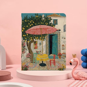 Puede incluir: Un cuaderno decorativo con una vibrante ilustración de una escena de café mediterráneo. La obra de arte representa una sombrilla roja sobre una mesa amarilla, una puerta turquesa y un limonero, creando un ambiente cálido. El cuaderno se exhibe sobre un pedestal rosa.