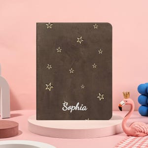 May include: A brown journal featuring gold star embellishments and the name "Sophia" in white script. The journal is displayed on a pink platform, with a pink flamingo ornament and blue sphere in the background.