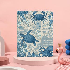 Puede incluir: Un diario azul y blanco con ilustraciones de criaturas marinas, como tortugas, cangrejos y medusas. El diario tiene el texto "GO WHERE YOU FEEL MOST ALIV". El diario está sobre una superficie rosa.