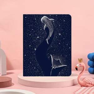 Może przedstawiać: Granatowy notatnik z białą ilustracją dwóch rekinów wielorybich pływających na gwiaździstym niebie. Projekt zawiera białe kropki reprezentujące gwiazdy i wzory rekinów wielorybich.