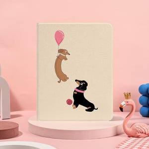 Könnte beinhalten: Eine beige Tablet-Hülle mit einer Illustration von zwei Dackeln. Einer schwebt mit einem rosa Ballon, der andere sitzt mit einem rosa Ball. Das Design ist verspielt und farbenfroh.