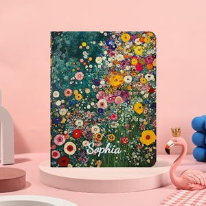 Op de afbeelding: Een notitieboek met een bloemmotief. De omslag toont een kleurrijk arrangement van bloemen in tinten geel, rood, roze en blauw tegen een teal achtergrond. De naam "Sophia" is in witte letters geschreven.
