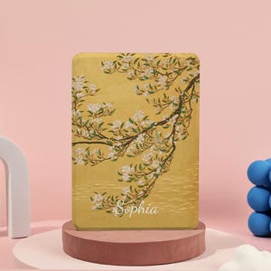 以下が含まれることがあります： 白い花と緑の葉が描かれた、花の枝のデザインが特徴の長方形の黄色いケース。「Sophia」という名前が筆記体で書かれています。ケースはピンク色の台座に展示されています。