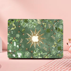 Puede incluir: Funda verde para portátil con diseño floral y de mariposas. La funda presenta un diseño de sol con el logotipo de Apple en el centro. La funda está decorada con hojas verdes, flores blancas y mariposas.
