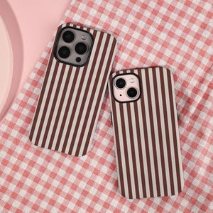 May include: Two phone cases with a vertical striped pattern in brown and cream. The cases are lying on a red and white checkered surface. The phone cases have camera cutouts.