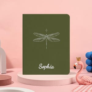 Puede incluir: Un cuaderno verde con una ilustración de libélula blanca y el nombre "Sophia" en cursiva. El cuaderno está sobre una plataforma rosa y blanca, con un flamenco rosa y una escultura de bolas azules en el fondo.