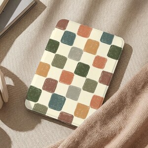 May include: A cream-colored tablet case featuring a repeating pattern of colorful, square shapes in shades of orange, green, blue, and brown. The case is resting on a neutral-toned surface, with a portion of a sweater visible.