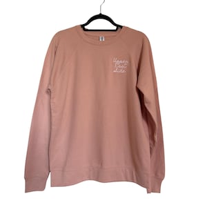 May include: A light pink sweatshirt with the words "Upper East Side" embroidered in white script on the upper left chest. The sweatshirt is hanging on a black hanger against a white background.