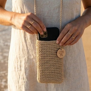 May include: A beige crochet crossbody phone pouch with a matching braided rope strap. The rectangular bag features a textured stitch pattern and a small round wooden tag attached to the front that reads Crochet Dreams with LOVE.