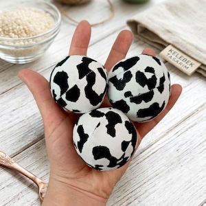 May include: Three white and black patterned balls are held in a hand. The balls have a cow-like pattern. Other items in the background include a small green plant, a bowl of seeds, and a roll of twine.