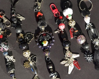 May include: A collection of Halloween-themed keychains with various charms. The keychains feature black beads, silver-tone metal charms, and decorative elements like ghosts, bats, coffins, and skulls. Each keychain has a different design and color accents, including red and white.