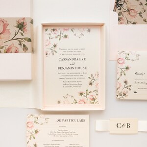 May include: A wedding invitation with a floral design featuring pink roses and green leaves. The invitation is printed on cream paper and includes the names Cassandra Eve and Benjamin House, the date and time of the wedding, and the location. The invitation is enclosed in a white envelope with a floral design.