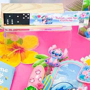 May include: A colourful party scene with a wooden domino set, character cutouts, and decorative elements. The scene includes a pink background, tropical flowers, and the name "Valentina" in a decorative sign. The theme appears to be a tropical or Hawaiian-themed party.