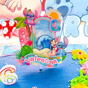 May include: A blue birthday cake decorated with a tropical theme. The cake features a topper with the name "Valentina", the number "6", and characters from the animated film Lilo & Stitch. The words "Happy Birthday" are also visible.